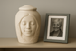 Personalized 3D memorial urn with a serene face design, created in resin to resemble a loved one, displayed beside a framed photo. Custom handmade urn by Always With Me.
